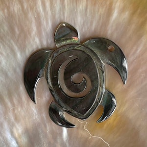 Peut inclure: Un pendentif en forme de tortue sculpté en noir avec un motif en spirale. Le pendentif est fait d'un matériau noir poli et a une finition lisse et brillante.