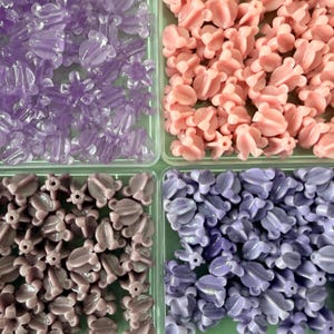 May include: An overhead shot of four compartments filled with decorative beads. The beads are in various shades of purple and pink. The beads are shaped like flowers and other organic shapes. The beads are made of a shiny material.