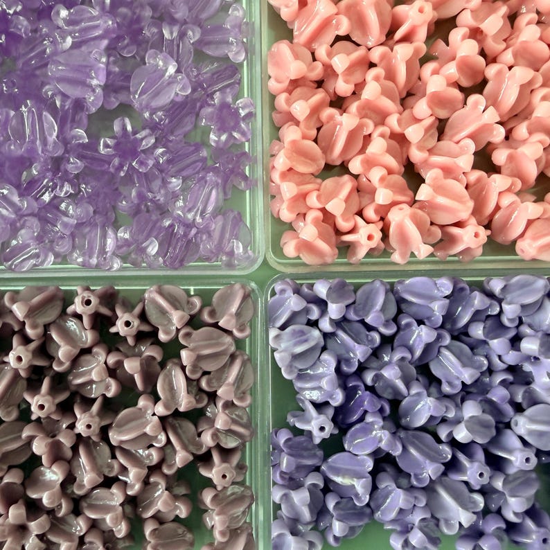 May include: An overhead shot of four compartments filled with decorative beads. The beads are in various shades of purple and pink. The beads are shaped like flowers and other organic shapes. The beads are made of a shiny material.