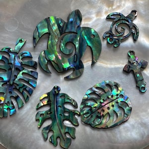 Può includere: Un set di sei ciondoli in conchiglia iridescente verde e blu con una tartaruga, una croce e quattro foglie di monstera.