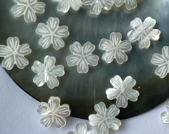 May include: A collection of small, white, flower-shaped mother-of-pearl beads. Each bead has five petals with etched details, scattered on a dark, iridescent surface. The beads are ideal for jewelry making or craft projects.