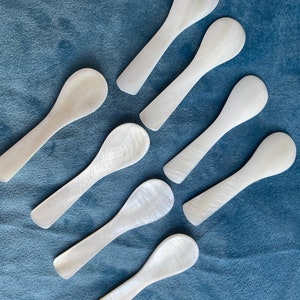 May include: Eight white mother-of-pearl spoons arranged in a pattern on a blue fabric background.