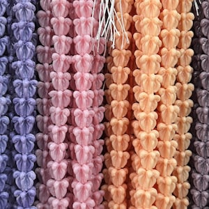 May include: Rows of colorful, decorative beads in various shades, including white, purple, pink, peach, and gray. Each bead is shaped like a stylized flower, strung together with white string, creating a textured, eye-catching display.