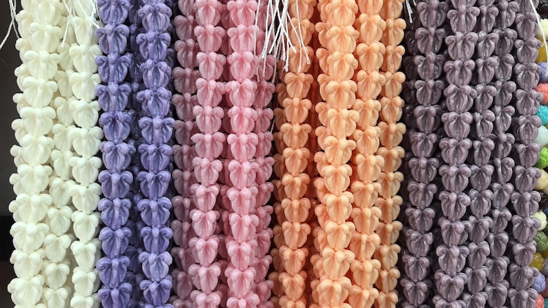 May include: Rows of colorful, decorative beads in various shades, including white, purple, pink, peach, and gray. Each bead is shaped like a stylized flower, strung together with white string, creating a textured, eye-catching display.