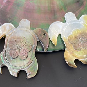 Puede incluir: Dos tallas de caparazón de tortuga de nácar iridiscente verde, marrón y blanco con un diseño de flor tallada en el centro.
