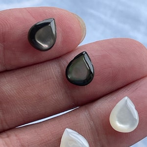 May include: Four teardrop-shaped cabochons with a smooth, polished finish. Two of the cabochons are a deep, iridescent charcoal black, while the other two are a creamy, pearlescent white. Each piece features a rounded top and a pointed base.