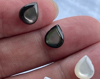 May include: Four teardrop-shaped cabochons with a smooth, polished finish. Two of the cabochons are a deep, iridescent charcoal black, while the other two are a creamy, pearlescent white. Each piece features a rounded top and a pointed base.