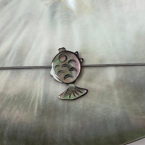 May include: A black and iridescent mother-of-pearl fish charm with a detailed design. The charm is on a silver wire.