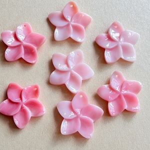 May include: Seven light pink flower-shaped charms arranged in a cluster. Each charm features five smooth, rounded petals and a small hole at the top for attachment. The material has a glossy, semi-translucent finish with a soft, uniform color.