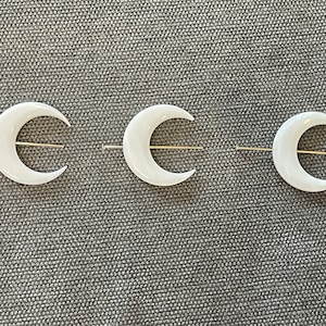 Pode incluir: Três grampos de cabelo em forma de lua crescente branca com barras de metal dourado. Os grampos estão dispostos em fila sobre uma superfície cinzenta texturizada.
