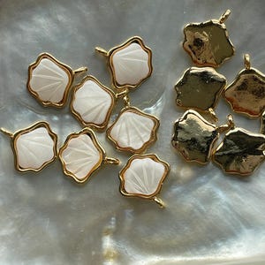 Puede incluir: Una colección de abalorios de metal dorado con un diseño de concha blanca. Los abalorios tienen un borde festoneado y un bucle para sujetarlos a joyas.
