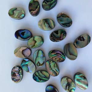 May include: A collection of oval-shaped abalone shell cabochons in various shades of green, blue, and iridescent colors. The cabochons are smooth and polished, with a natural, organic look.