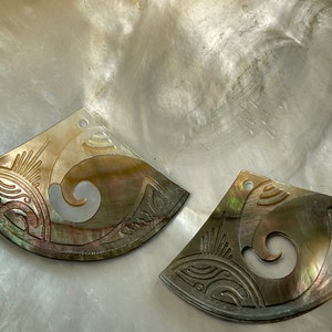 Könnte beinhalten: Zwei irisierende Perlmutt-Anhänger mit aufwendigen Schnitzereien. Die Anhänger sind halbmondförmig und haben ein Spiralmuster.