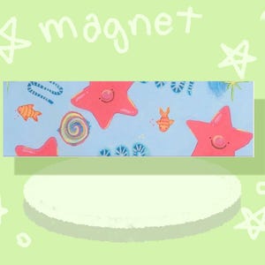 May include: A rectangular magnet featuring a light blue background with a pattern of pink starfish, orange fish, and colorful spiral shapes. The word magnet is written in white text above the rectangular object, surrounded by white star doodles.