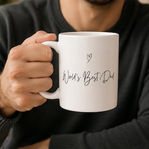 Pode incluir: Uma caneca de cerâmica branca com um ícone de coração desenhado à mão em preto acima do texto cursivo preto que diz World's Best Dad. A caneca tem um corpo cilíndrico liso e uma alça curva na lateral, oferecendo um design simples e limpo.