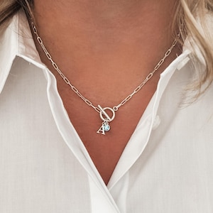 Könnte beinhalten: Eine silberne Kettenkette mit einem Toggle-Verschluss und einem kleinen Anhänger mit dem Buchstaben 'A' und einem blauen Edelstein.