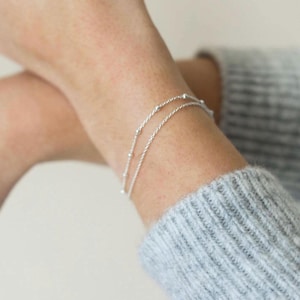Könnte beinhalten: Ein silbernes Kettenarmband mit kleinen Perlen. Das Armband besteht aus zwei übereinanderliegenden Ketten.