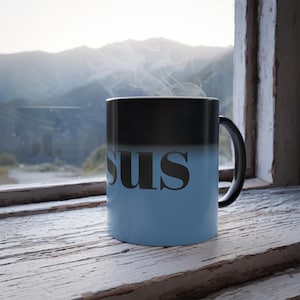 May include: A black and blue color-changing mug with the word "sus" printed in black. The mug is sitting on a weathered, white wooden windowsill. Steam rises from the mug, suggesting a hot beverage.