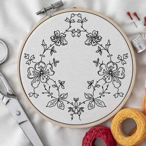 Pode incluir: Um bastidor de bordado circular de madeira contendo tecido branco com um padrão floral de linhas pretas. O design apresenta caules, folhas e flores simétricas dispostas em forma de coroa. Tesouras de metal, um dedal e fio estão próximos.