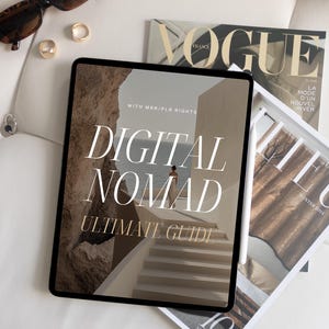 May include: A digital tablet displaying a guide titled "Digital Nomad Ultimate Guide" with a woman walking on a path in the background. The tablet is on a white surface with a magazine titled "Vogue" and another magazine titled "Lieu" visible in the background.
