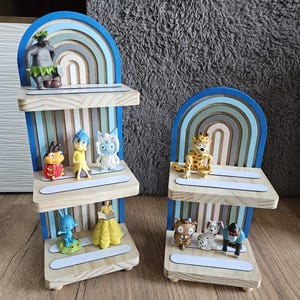 Puede incluir: Dos estanterías de madera con un diseño de arco iris en azul, gris y blanco. Cada estantería contiene varias figuras, incluyendo un gorila, un conejo azul y una figura de vestido amarillo. Las estanterías están sobre un suelo de madera.