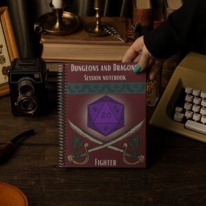 May include: A spiral-bound "Dungeons and Dragons" session notebook with a purple die and crossed swords graphic. The notebook is held by a person with teal nail polish. Other items include a vintage camera, typewriter, and framed mushroom print.