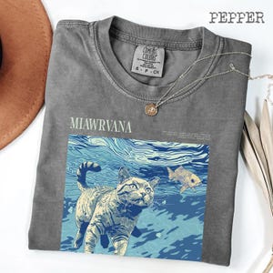May include: A gray Comfort Colors t-shirt with the text "MIAWRVANA" and a graphic of a cat underwater with a fish. A silver necklace with a pendant is draped across the shirt.