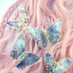 May include: Four iridescent blue butterfly hair clips with gold accents and rhinestones on a pink wig.