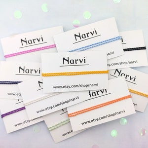 Può includere: Una collezione di braccialetti dell'amicizia colorati con la parola "Narvi" e l'indirizzo del sito web "www.etsy.com/shop/narvi" stampati su cartoncini bianchi.