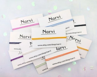 Può includere: Una collezione di braccialetti dell'amicizia colorati con la parola "Narvi" e l'indirizzo del sito web "www.etsy.com/shop/narvi" stampati su cartoncini bianchi.