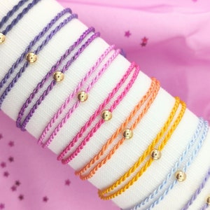 Può includere: Un set di braccialetti intrecciati colorati con perline dorate. I braccialetti sono disposti a forma di arcobaleno, con colori che vanno dal viola al blu, dal giallo all'arancione e al rosa. Tutti i braccialetti sono realizzati con lo stesso tipo di cordoncino e sono tutti legati con un semplice nodo.