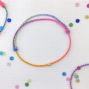 May include: Three colourful friendship bracelets with gold beads. The bracelets are made with a variety of colours, including pink, blue, green, and yellow. The bracelets are tied in a knot at the ends.