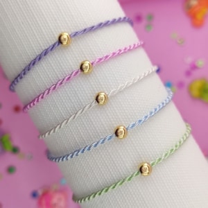 May include: Five colorful braided bracelets with gold beads. The bracelets are layered on a white surface. The bracelets are in shades of purple, pink, white, blue, and green.