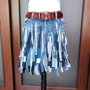 Op de afbeelding: Een spijkerbroekrok met een gefranjerde zoom, gemaakt van stroken denim in verschillende blauwtinten. Het wordt gedragen met een bruine leren riem met een gouden gesp. Een spijkerbroek crop top is ook zichtbaar.