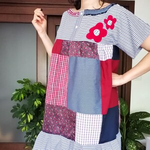 Può includere: Un abito patchwork con un mix di fantasie e tessuti, tra cui righe, quadri e stampe floreali. L'abito ha maniche corte, un orlo a balze ed è decorato con due applicazioni di fiori rossi. La palette di colori comprende rosso, bianco, blu e blu navy.