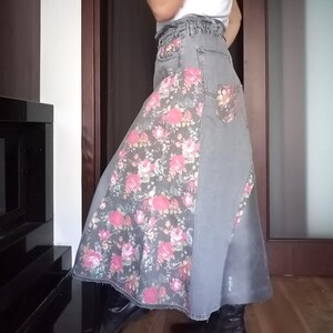 Op de afbeelding: Een lange spijkerbroekrok met een bloemenpatroon in roze en wit. De rok is lichtgrijs van kleur en heeft een hoge taille. Het bloemenpatroon staat op de voorkant en op de achterzak.