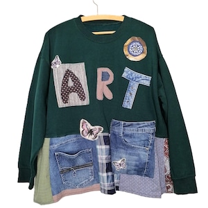 May include: A dark green sweatshirt with the word "ART" appliqued on the front. Patches of denim, patterned fabric, and butterfly appliques are sewn onto the sweatshirt, creating a unique, artistic design.