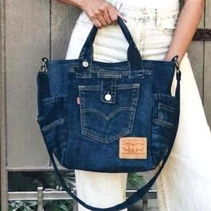Può includere: Una borsa tote in denim blu scuro con una tasca frontale con chiusura a bottone, tasche laterali e una toppa color cuoio sul fondo. La borsa include due manici superiori e una lunga tracolla regolabile realizzata in tessuto denim abbinato.