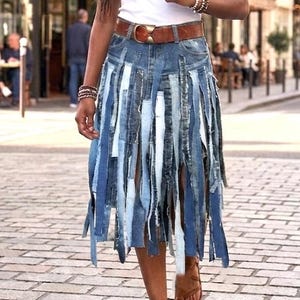 May include: A white tank top paired with a knee-length denim skirt featuring vertical strips of various blue-toned denim fabrics. The skirt is secured with a brown leather belt featuring a gold-toned buckle, and brown strappy leather sandals.