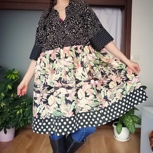 Op de afbeelding: Een patroonjurk met een zwart-witte top, een bloemenmiddenstuk en een zoom met stippen. De jurk heeft korte mouwen en een V-hals. Het model draagt een blauwe spijkerbroek en zwarte laarzen.