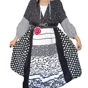 Puede incluir: Un vestido patchwork con falda de lunares blancos y negros, paneles florales y a rayas, y una parte superior negra con estampado floral. El vestido se ciñe a la cintura con un cinturón negro y un adorno floral rojo.