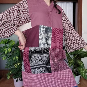 May include: A patchwork tunic in shades of burgundy, mauve, and white. The tunic features a V-neck, long sleeves with a floral pattern, and a mix of patterned and solid fabric panels. The hemline has a small polka dot pattern.