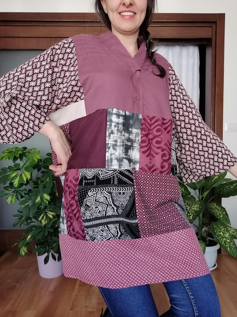 May include: A patchwork tunic in shades of burgundy, mauve, and white. The tunic features a V-neck, long sleeves with a floral pattern, and a mix of patterned and solid fabric panels. The hemline has a small polka dot pattern.