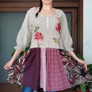 Op de afbeelding: Een beige tuniek met bloemenborduurwerk en een patchwork rok. De top heeft rode klaproosborduurwerk en driekwart mouwen. De rok is een mix van stoffen met patronen in bordeauxrood, rood en wit. De outfit wordt gedragen met een blauwe jeans.