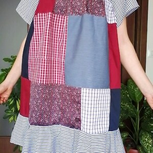 May include: A patchwork dress in red, blue, and white, with short sleeves and a ruffled hem. The dress incorporates striped, checked, and floral patterned fabrics. It features a red flower applique. The dress is a unique, handcrafted design.