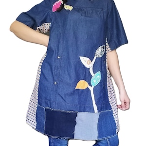 Pode incluir: Um vestido túnica de ganga com mangas curtas e um design de patchwork. O vestido apresenta um caule branco com aplicações de folhas coloridas. A bainha é um patchwork de quadrados de ganga. O vestido é combinado com jeans azuis e botas pretas.