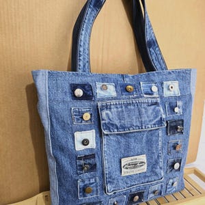 Puede incluir: Un bolso tote de mezclilla azul con un diseño de patchwork. La bolsa presenta varios parches de mezclilla, botones y un bolsillo frontal. Las asas están hechas del mismo material de mezclilla. Una etiqueta en la parte delantera dice "Better Jeans".