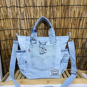Op de afbeelding: Een lichtblauwe denim tote bag met twee handvatten en een afneembare schouderriem. De tas heeft een centraal vak met een knoopsluiting, een klein rood Levi's-merklabel en een rechthoekige witte patch met zwarte tekst en afbeeldingen.