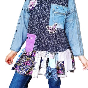 Op de afbeelding: Een tuniek met lange mouwen, een donkerblauw bloemenpatroon op het lijfje, lichtblauwe spijkerstof mouwen en een patchwork zoom. Het kledingstuk heeft een paarse zak, een vlinderapplicatie, een borstzak met rits en een knoopsluiting aan de voorzijde.