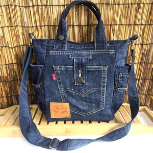 May include: A dark blue denim tote bag featuring two top handles and a detachable shoulder strap. The bag includes a central pocket with a button closure, a small red tag, and a tan leather-coloured patch with visible text and graphics.
